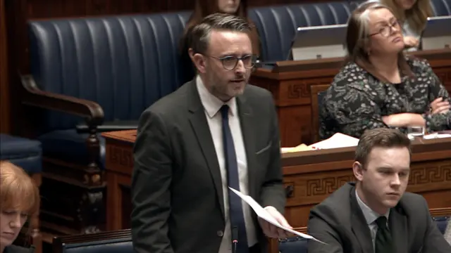 Nick Mathison standing in the Assembly chamber. He is wearing a dark grey suit, white shirt, navy tie and glasses.