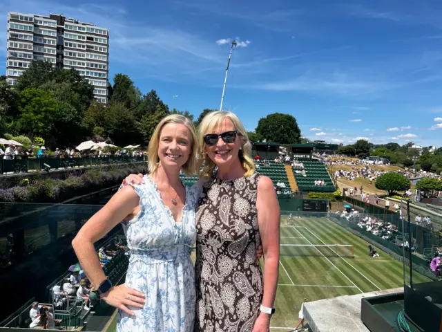 Carol and Sarah smiling together at a tennis stadium, standing by the court with spectators, greenery, and a tall apartment building