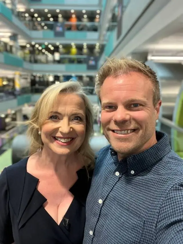 Carol and Simon smile for a selfie inside London Broadcasting House, with lighting, glass railings, and people visible on floors behind them.