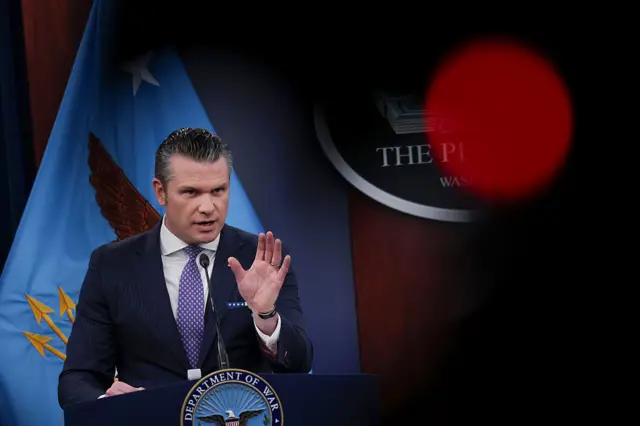 US Defence Secretary Pete Hegseth with one hand raised and looking across the room seriously. There is a flag with an eagle behind him, and a shadow of a person's head in the foreground