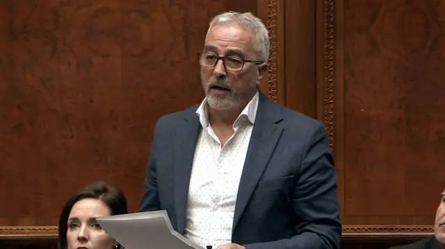 Pat Sheehan, with short grey hair, wearing a white shirt, navy suit and glasses. He is standing in the Assembly chamber.