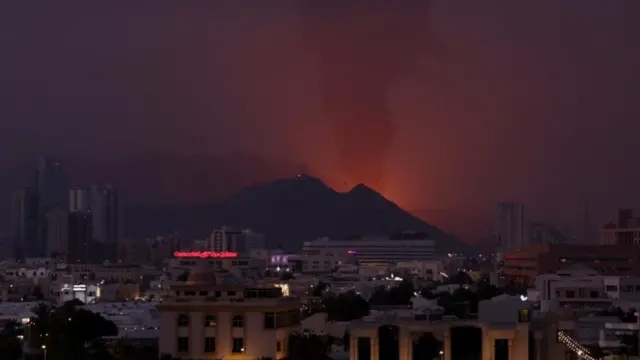 red smoke rising over a pink sky above mountains with city in foreground