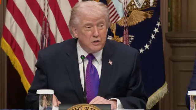 President Trump, wearing suit and purple tie, speaking into a microphone at a table with an American flag behind him