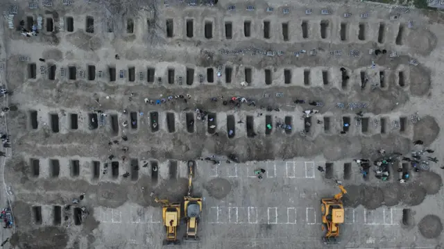 Three yellow construction vehicles are seen digging rectangular grave sites, which have been marked out by white chalk.