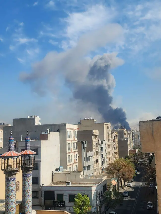 A plume of smoke rising several streets away from the picture, in the foreground two turquoise and blue minarets can be seen