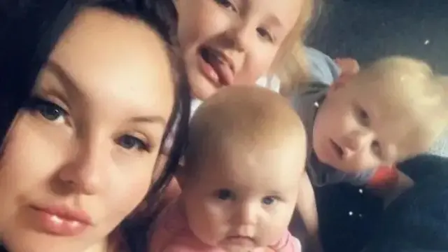 A brown-haired young woman next to three young blonde-haired children. They are all staring directly into the camera.