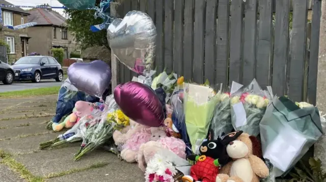 A floral tribute featuring bunches of flowers, heart-shaped balloons and cuddly toys. They are resting against a grey garden fence on a residential street.