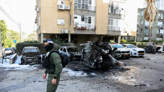 A khaki-dressed officer wearing a police balaclava walks past a blackened charred cars, one of which is flipped on its side.
