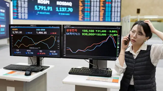 A woman in a black puffer vest scratches her head while speaking over the phone. She is standing in front of an electronic scoreboard at the Korea Exchange in Seoul, South Korea. The screens display sharply fallen closing indices.
