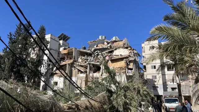 Wide shot of a heavily damaged building in Beirut, the top floors are collapsed onto each other. On the bottom right side of the picture are at least six men observing the scene near a white car