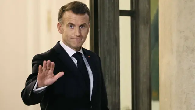 Emmanuel Macron holds his hand out to the camera. He wears a black suit jacket and tie
