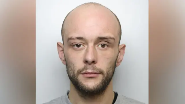 A bald man in his 20s staring directly into the camera. He has brown eyes, a short beard and a grey jumper.