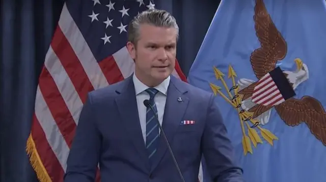 Америкийн далбааны өмнө микрофонд ярьж буй Хгсет