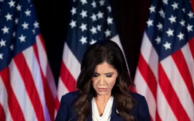 Kristi Noem looks down while standing in front of a background of flags
