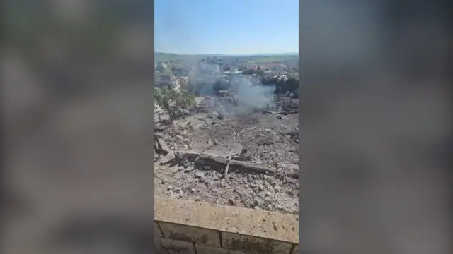 A screenshot of footage shows a smouldering wreckage in front of several rural buildings