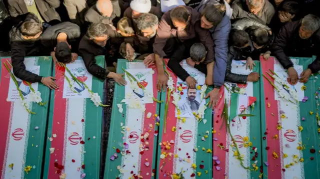 Mourners plan their hands on coffins which have been covered with the flag of Iran. The coffins have been decorated with flowers and pettles