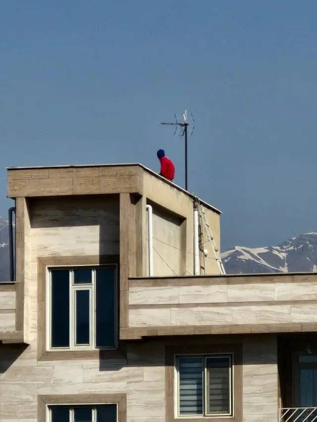 Man in red jumper sits on top of a building, mountains can be seen in the background.