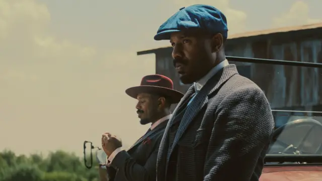 Michael B Jordan playing twins Smoke and Stack, his two characters both wear suits, one has a fedora on, the other is wearing a baker boy cap