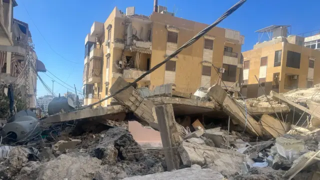 Wide shot showing debris of buildings in Saida, Lebanon. There's three residential four-storey buildings in the background showing signs of heavy damage