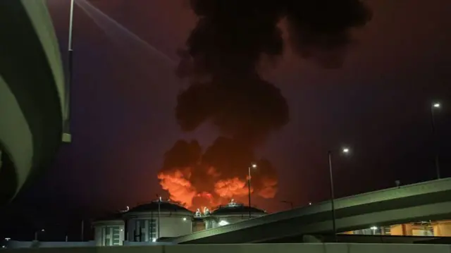 black and orange smoke rising from large cylindrical fuel containers behind a freeway