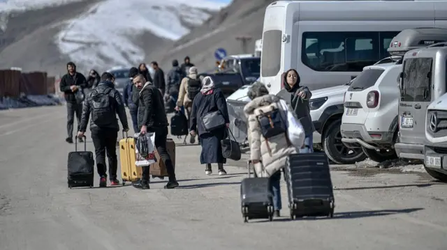 People wearing winter clothing carry suitcases along a road with cars beside and snow capped mountains in the distance