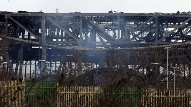 The metal structure of a sports stadium is exposed following a strike. Smoke rises in front of the damaged building