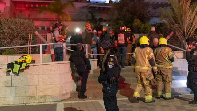 Israeli police and firefighters crowd outside a building hit by a missile in Tel Aviv. A female police officer stands at the front looking at the ground.