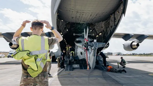A man in a high-vis vest and military fatigues stands in the foreground as a Wildcat helicopter is unloaded from the back of a C-17