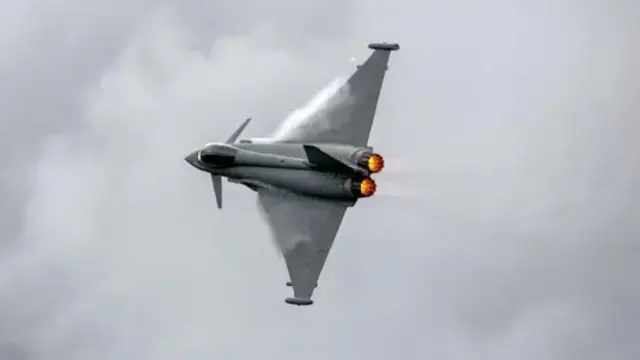 A grey aircraft with long wings and fire at its tail in front of a grey sky covered by white clouds.
