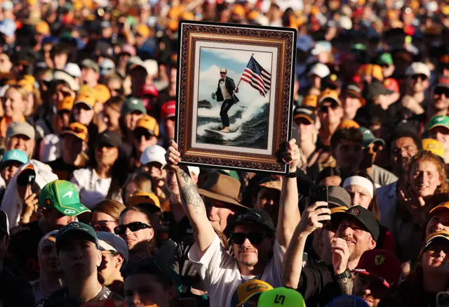 A fan holds up a poster of Valtteri Bottas on a surfboard, wearing a suit and holding an American flag