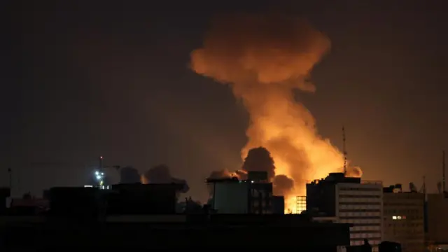 A dark sky with orange and white smoke rising between buildings and into the sky.