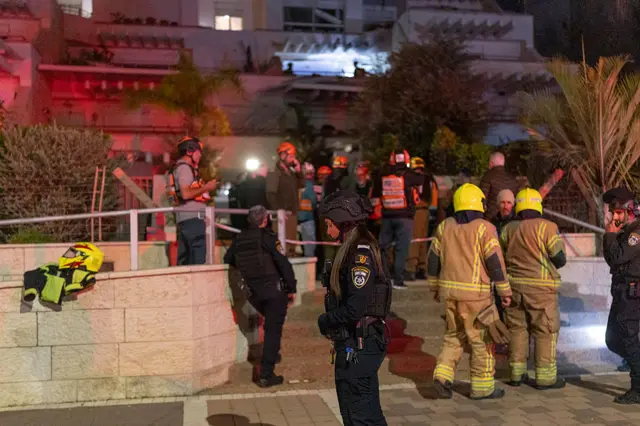 Israeli police and firefighters crowd outside a building hit by a missile in Tel Aviv. A female police officer stands at the front looking at the ground.
