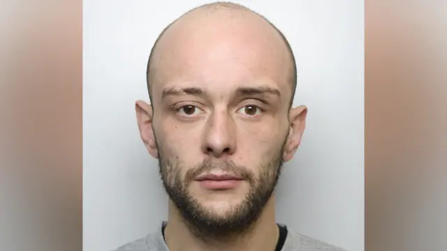 A police mugshot of Calum Sunderland. He has a bald head and a dark beard. He is wearing a grey jumper.