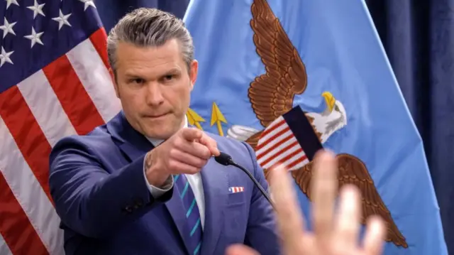 US Secretary of Defense Pete Hegseth points to a person in the crowd while standing in front of the American flag and the Great Seal of the United States on a blue flag.