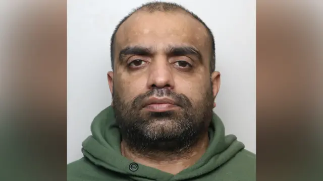 A police mugshot of Sharaz Ali. He has short dark hair and a dark beard. He is wearing a green hooded top.