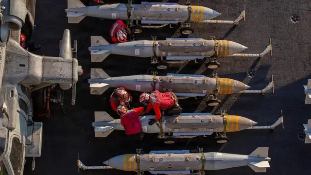 A bird's eye view of a stockpile of missiles in a line with US Navy commanders working on them in red suits.