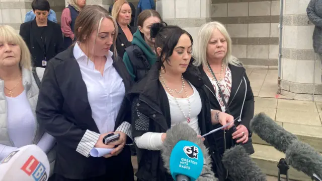 A group of women stand outside in front of a collection of microphones. The woman at the centre is reading from a piece of paper