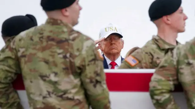 Trump, wearting a white baseball hat that reads "USA," closes his eyes as soldiers carry a casket in front of him as he salutes