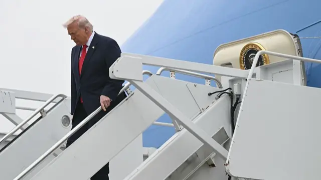 Donald Trump holds a silver railing as he walks down the steps of Air Force one with his eyes closed and his head tilted downwards, he is wearing a navy suit and red tie