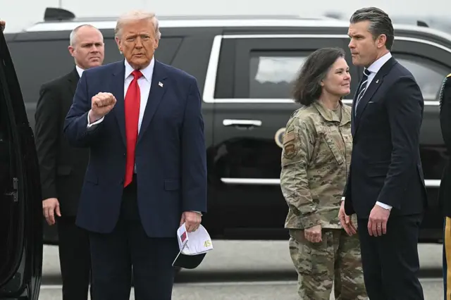 Donald Trump holds his right first up while holding a white baseball hat in his left hand, Defence Secretary Pete Hegseth stands nearby as do other members of the military