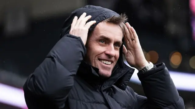 A smiing Ipswich Town head coach Kieran McKenna putting the hood of his coat up over his head