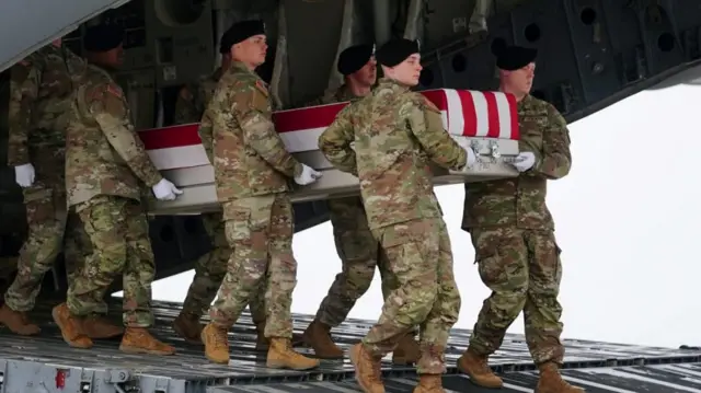 Soldiers in green and brown uniforms carry a casket drapped with an American flag off of a plane