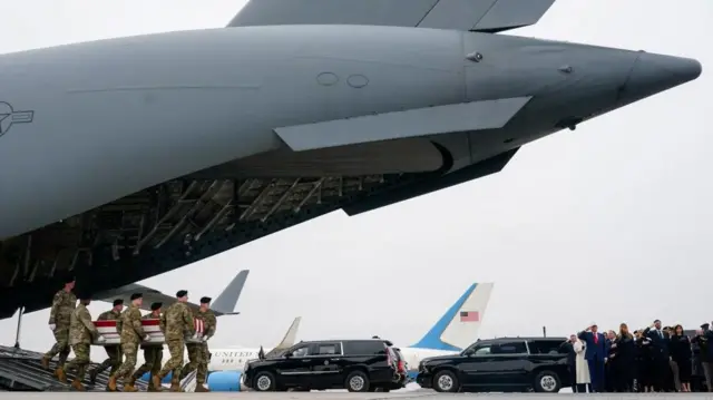 A wide shot showing the tail end of a military plane as soldiers carry a casket off of it and members of the Trump administration salute
