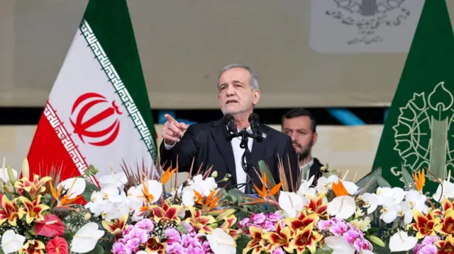 Pezeshkian stands in front of Iranian flags and behind a table full of flowers.