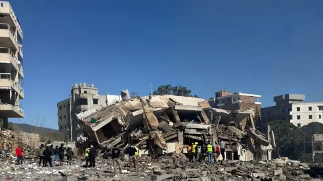 Rescue workers stand among debris in front of a collapsed multi‑storey building, surrounded by damaged structures, scattered rubble and a bright blue sky overhead