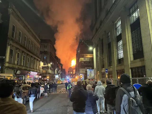 Night sky lit up with flames. Lots of people in the street watching.