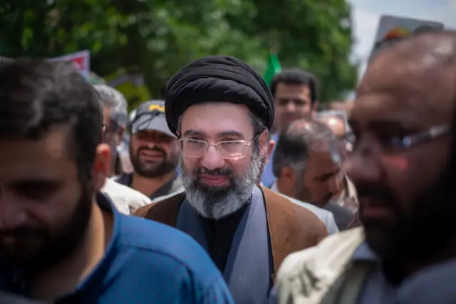 Mojtaba Khamenei in cleric dress with brown cloak and black head cover walks in a crowd