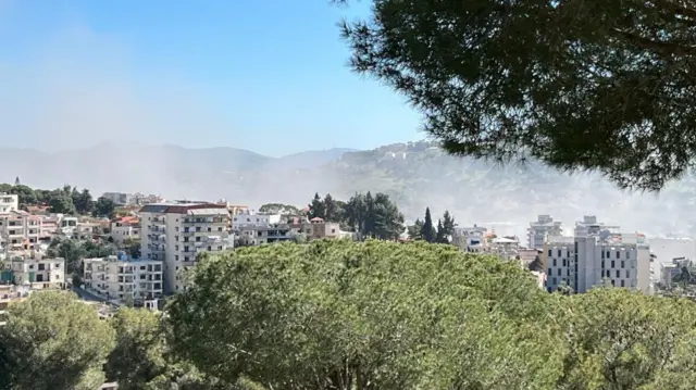 Wide view of a hillside town with apartment buildings and trees in the foreground, as pale smoke or haze drifts across the valley under a clear blue sky