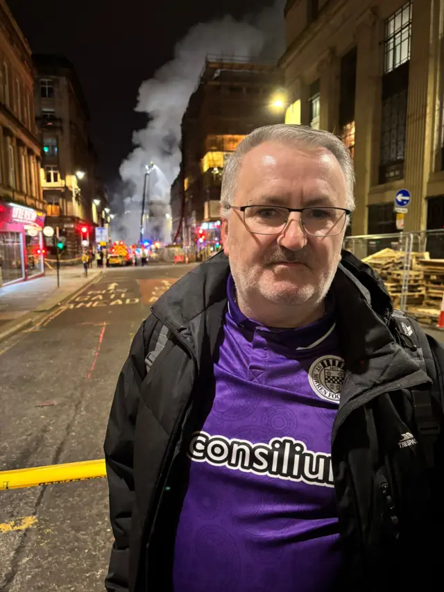 A grey-haired man in a purple St Mirren football shirt, standing in front the Glasgow fire