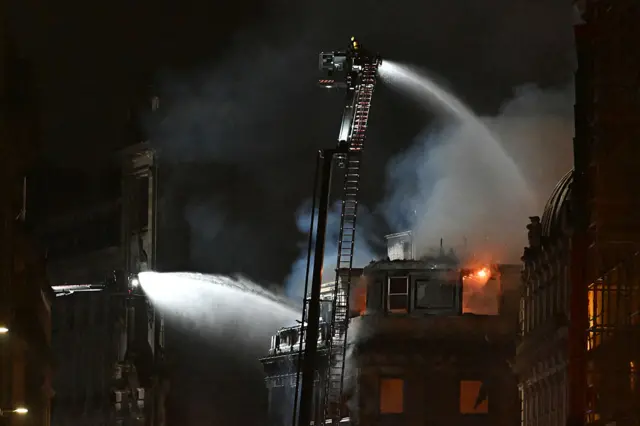 Night-time image of firefighters on a tall ladder, spraying water into a burning building